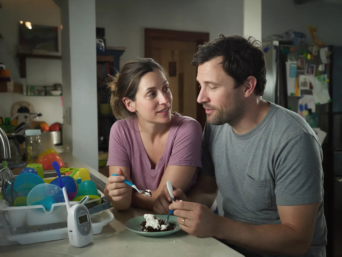 A couple sharing a dessert at the kitchen counter beside a baby monitor and bottles